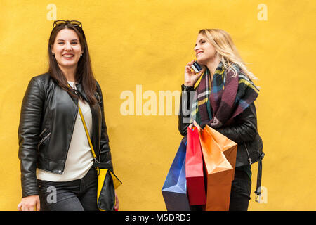 Zwei nette Mädchen brünetten und blonden haben einem Shopping Tag am schwarzen Freitag. Einer von ihnen im Gespräch mit einem Telefon andere lächelt. Vor eine einfache gelbe Wand Stockfoto