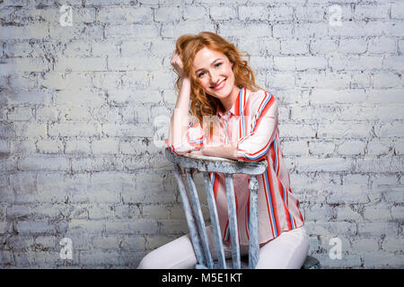 - Nahaufnahme portrait Schöne junge Studentin mit rotem lockigem Haar und Sommersprossen im Gesicht sitzen auf einer Mauer in grauen Hintergrund. In einem gekleidet Stockfoto