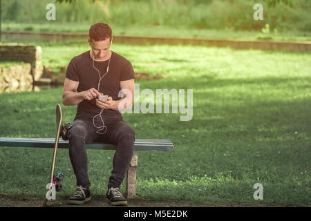 Urban Mann sitzt auf der Bank im Park und mit Musik App auf sein Smartphone. Sein Skateboard ist neben ihm. Stockfoto