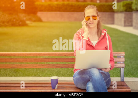 Urban casual Frau sitzt auf einer Bank im Park. Sie ist Gespräch am Handy und Laptop. Tasse Kaffee zu gehen ist neben ihr. Stockfoto