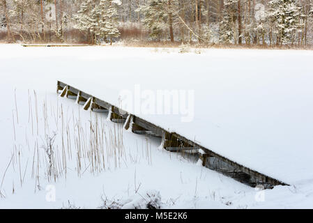 Kleine und schmale hölzerne Seebrücke im winterlichen Wald See, Schnee und Eis auf den See und die umliegende Landschaft. Stockfoto