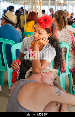 Ein kaukasischer Mann und seiner philippinischen Begleiter Streik eine lustige darstellen, da sie erschöpft Sitzen beim Warten auf die Fähre in Puerto Princesa, Palawan, Philippinen Stockfoto