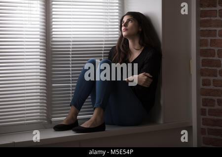 Nachdenkliche junge Frau sitzt auf der Fensterbank Stockfoto