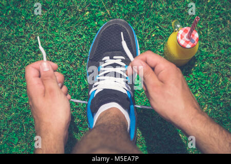 Hohe Betrachtungswinkel Nahaufnahme von jogger Schnürsenkel binden auf Laufschuhe im Feld und Flasche Glas Orangensaft mit Trinkhalm. Sport und heilen Stockfoto