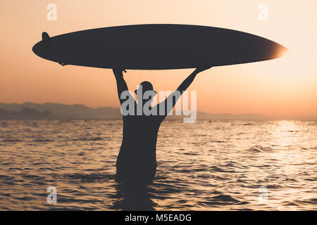 Touristische Kerl in Hut stehend im Wasser und dem Surfboard bei Sonnenuntergang. Freiheit und Sommerurlaub Konzepte. Stockfoto