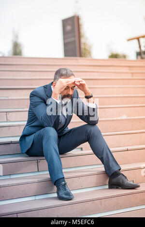 Gestresst und überfordert älterer Geschäftsmann versucht, die Spannung zu lösen, sitzen auf der Treppe in einem städtischen Außenbereich Stockfoto