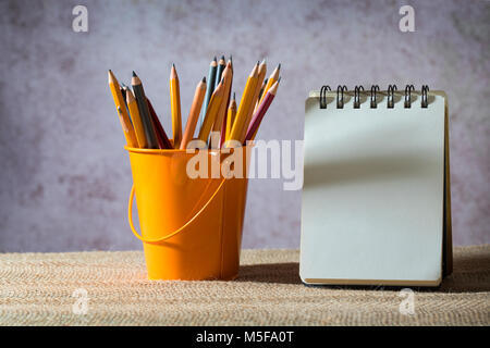 Eine Reihe von Buntstiften auf ein Spielzeug Eimer und ein leeres kleines Notebook Stockfoto