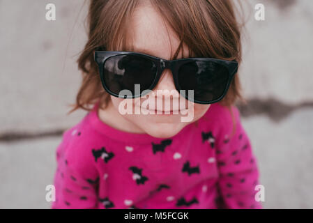 Ein Kleinkind Mädchen mit einem rosa Hemd und Sonnenbrille, stehend auf einem Bürgersteig an einem warmen, sonnigen Tag. Stockfoto