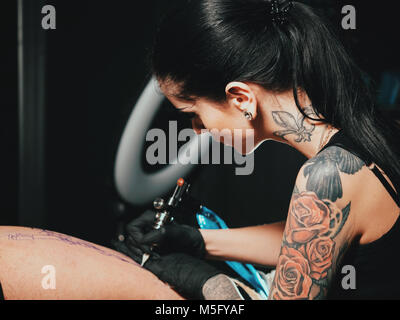 Close up Tattoo artist demonstriert den Prozess des Erhaltens Schwarz Tattoo mit Farbe. Konzentriert weiblichen Färbung Kontur mit schwarzer Tinte auf Bein mit gu Stockfoto