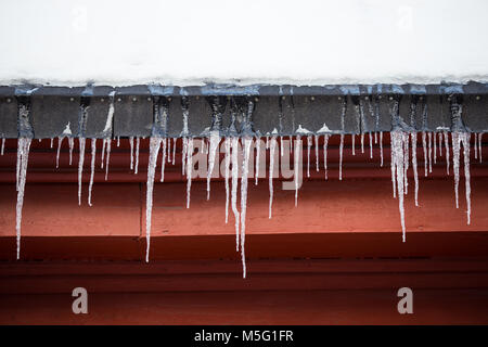 Detail von Eiszapfen hängen von Schnee bedeckte Dach auf roten Gebäude aus Holz Stockfoto