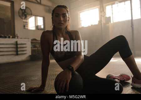 Junge Frau sitzt auf der Turnhalle Boden mit medizinball nach der Trainingseinheit. Weibliche eine Pause vom Training. Stockfoto