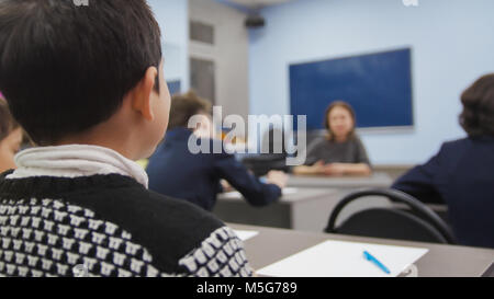 Klassenkameraden auf Schule - Junge sitzt am Tisch und Suchen während der Lehrer erklärt die Lektion Stockfoto