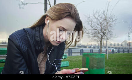 Enttäuscht Mädchen mit langen blonden Haaren in Leder Jacke begradigt verwenden Gadget auf der Bank sitzen im Wind Kopfhörer anhören, close-up Stockfoto