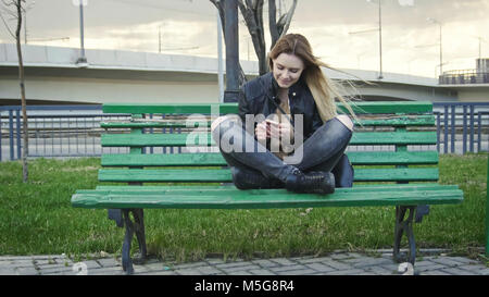 Cute glücklich Mädchen mit langen blonden Haaren in Leder Jacke begradigt verwenden Gadget auf der Bank sitzen im Wind, horizontal Stockfoto