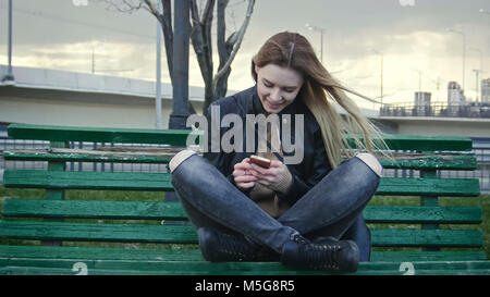 Cute glücklich Mädchen mit langen blonden Haaren in Leder Jacke begradigt verwenden Gadget auf der Bank sitzen im Wind Stockfoto