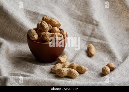 Zusammensetzung der rohe ungeschälte Erdnüsse in Ton Keramik Schüssel auf hellgrau Baumwolle Tischdecke, Servietten, etwas Platz, closeup kopieren, selektiven Fokus. Gesund Stockfoto