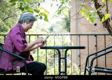 Asiatische ältere Frau mit Mobiltelefon auf der Terrasse. ältere weibliche sms Nachricht, mit App mit Smartphone auf Terrasse. Senior mit Handys zu verbinden Stockfoto