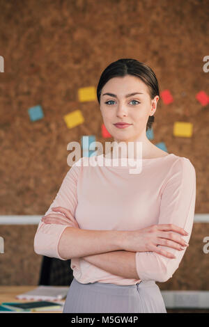 Portrait einer weiblichen Büroangestellten. Sie steht vor einem Cork board, elegant gekleidet mit ihren Armen gefaltet und ist ein Lächeln für die Kamera. Stockfoto