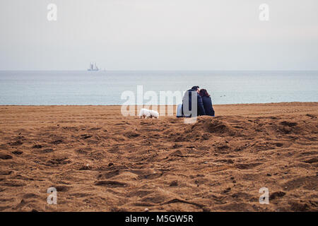 BARCELONA, SPANIEN - 17. FEBRUAR 2018: Сouple der Liebhaber mit einem Hund küssen Auf den Sant Miguel Strand mit einem Segelschiff am Horizont Stockfoto