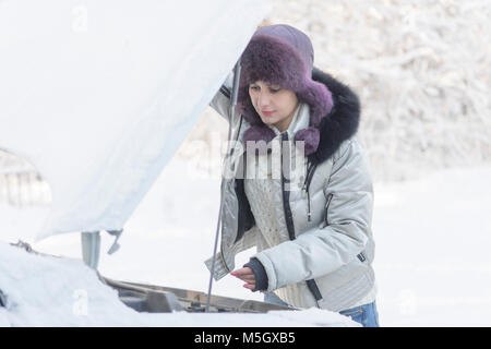 Das Mädchen hob die Haube vom Auto und schaut auf den Motor. Winter, Tag. Stockfoto