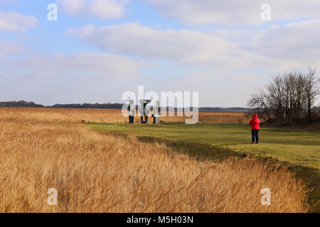 Snape Maltings, Suffolk. 23 Feb, 2018. UK Wetter: eine Dame in einem roten Mantel und Hut Studien der Familie der Mann Skulptur von Barbara Hepworth auf einem hellen sonnigen Nachmittag auf Snape Maltings, Suffolk. Stockfoto