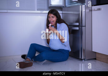 Junge Frau sitzt auf dem Boden essen Kuchen in der Küche Stockfoto