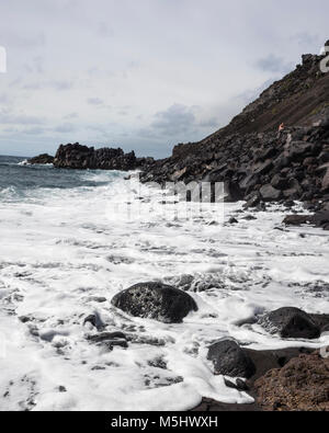 Salinas de Fuencaliente, La Palma. Kanarische Inseln Spanien. Viele Sea Foam ist vom Meer Wasser hetzen durch die lavafelsen an einem kleinen Strand. Dieses kleine Fuencaliente de La Palma Bay ist typisch für die dramatische Küstenlinie der Insel. Die lavafelsen steigen stark an eine Felswand zu bilden. Die Sonne scheint hell die Sea Foam sehr weiß und der Himmel Hellblau. Die großen lavafelsen im Vordergrund durch das Meer, das die Härte der Meeresumwelt erodiert. Mit einer Ricoh GRII Kamera fotografiert. Stockfoto