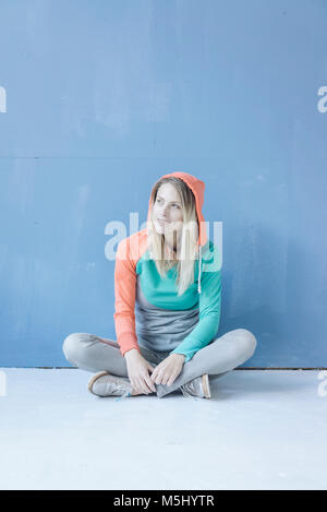 Porträt der lächelnde Frau tragen Kapuzenjacke sitzen auf dem Boden vor der blauen Wand Stockfoto