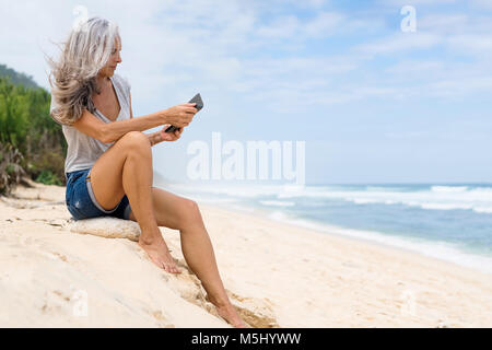 Schönen Lächeln ältere Frau am Strand sitzen, Holding e-book Stockfoto