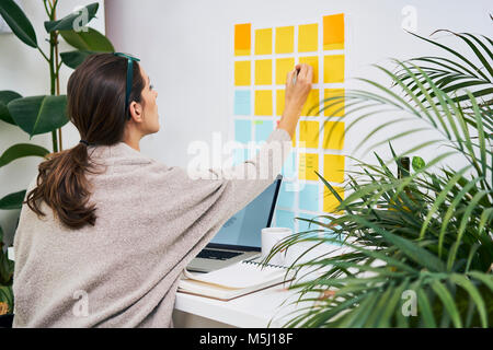 Junge Frau mit Laptop auf dem Schreibtisch arbeiten mit Haftnotizen an der Wand Stockfoto