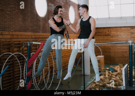 Zwei lächelnde Gymnasts sitzen auf hohen bar Händeschütteln in der Turnhalle Stockfoto