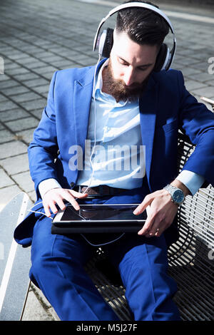 Junger Mann am Bahnhof sitzen, mit digitalen Tablet und Kopfhörer Stockfoto