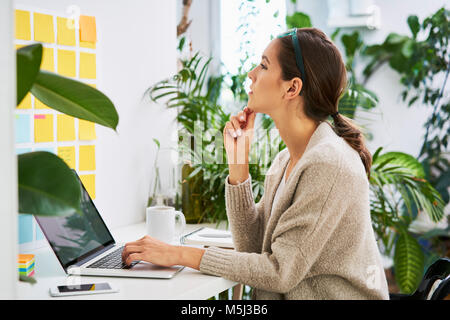 Junge Frau mit Laptop auf dem Schreibtisch auf der Suche nach Haftnotizen an der Wand Stockfoto