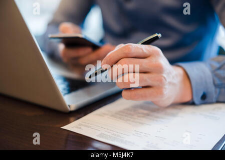 Mann an Schreibtisch mit Handy und Kugelschreiber in der Hand, Nahaufnahme Stockfoto