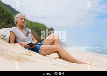 Schönen Lächeln ältere Frau am Strand liegend, Holding e-book Stockfoto
