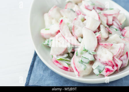 Gesunden roten Rettich und Grünen Schalotten Zwiebeln Salat mit griechischen Joghurt Dressing in Weiß Servierschale stehend auf Blau Serviette. Osterbrunch Mahlzeit Feder Stockfoto