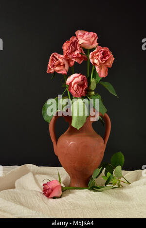 Fünf sterben rosa Rosen in einer Vase Terrakotta vor einem schwarzen Hintergrund, mit einer einzelnen Rose auf musselin Tuch. Stockfoto
