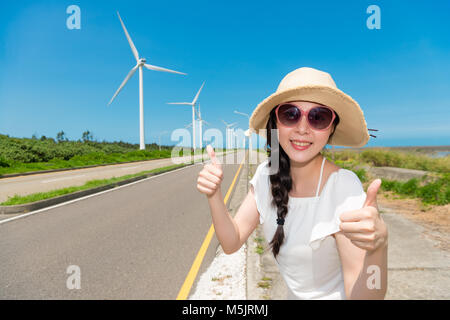 Gerne asiatische Frau macht einen Daumen nach oben, zu lieben und sich auf Reisen mit grünen Windmühle Power für Ihren Urlaub mit tollen, sonnigen Wetter Stockfoto