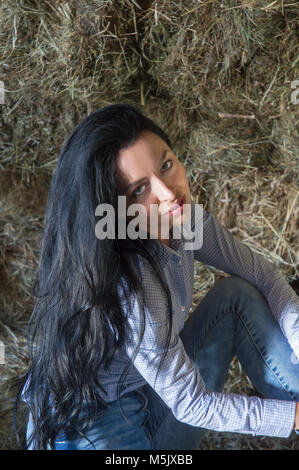 Lange schwarze Haare weiblichen Blick auf Kamera, während in der Scheune sitzen Stockfoto