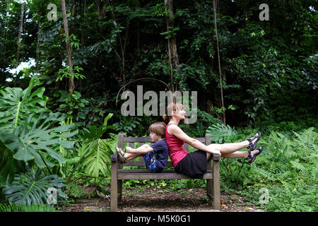 Mutter und Sohn verspielter sitzen Rücken an Rücken auf einer Holzbank im tropischen Garten der Fort Canning Park, Singapur Stockfoto