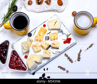 Kleine Stücke von Brie Käse, Roquefort, Camembert, Cheddar und Käse mit Walnüssen auf ein weißes Holzbrett, neben zwei gelb Keramik Becher mit Schwarz Stockfoto