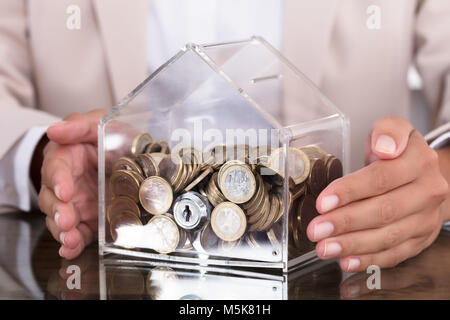 Mittelteil der Geschäftsfrau Schutz der Münzen in transparenten Haus geformte Sparschwein an den hölzernen Tisch Stockfoto