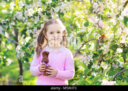 Kinder mit Hasenohren auf Ostereiersuche im Blooming Cherry Blossom Garten. Kleines Mädchen Schokolade essen Kaninchen. Frühling Blumen und Eier Warenkorb in frui Stockfoto