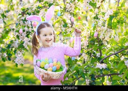 Kind auf Ostereiersuche im blühenden Kirschbaum Garten mit Blumen. Kind mit farbigen Eier im Korb. Kleines Mädchen mit Hasenohren. Ostern deco Stockfoto