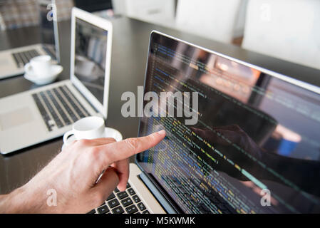 Mehrere Computer mit Kaffee in einer Büroumgebung mit Hand zeigen auf Bildschirm mit Zeilen Code. Stockfoto