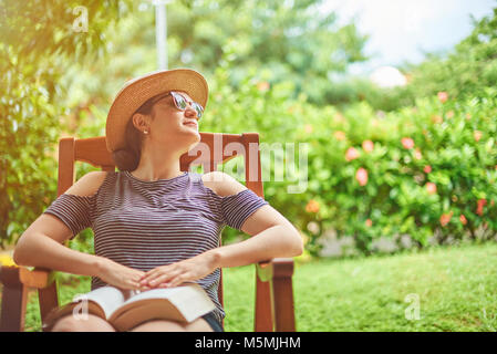 Junge Frau mit Buch sitzen auf Stuhl im grünen Garten Terrasse Stockfoto