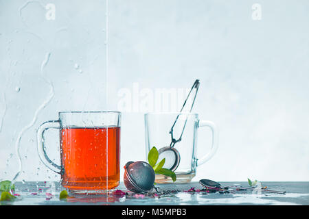 Tasse Tee und Tasse mit Teesieb, transparent auf einem hellen Hintergrund mit grünen Blättern. Der Frühling noch Leben mit Regentropfen auf dem Fenster. Stiftleiste mit kopieren. Stockfoto