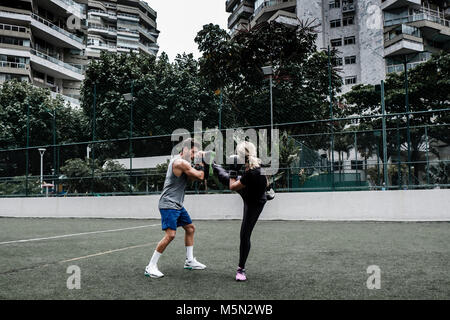 Erwachsene Frau training Muay Thai mit männlichen Trainer im grünen Feld mit Gebäude im Hintergrund Stockfoto