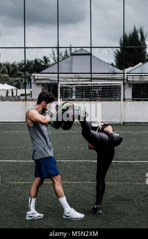 Erwachsene Frau training Muay Thai mit männlichen Trainer im grünen Feld mit Gebäude im Hintergrund Stockfoto