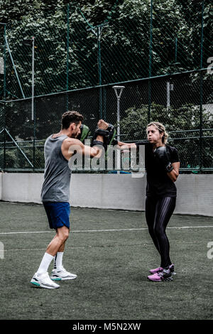 Erwachsene Frau training Muay Thai mit männlichen Trainer im grünen Feld mit Gebäude im Hintergrund Stockfoto